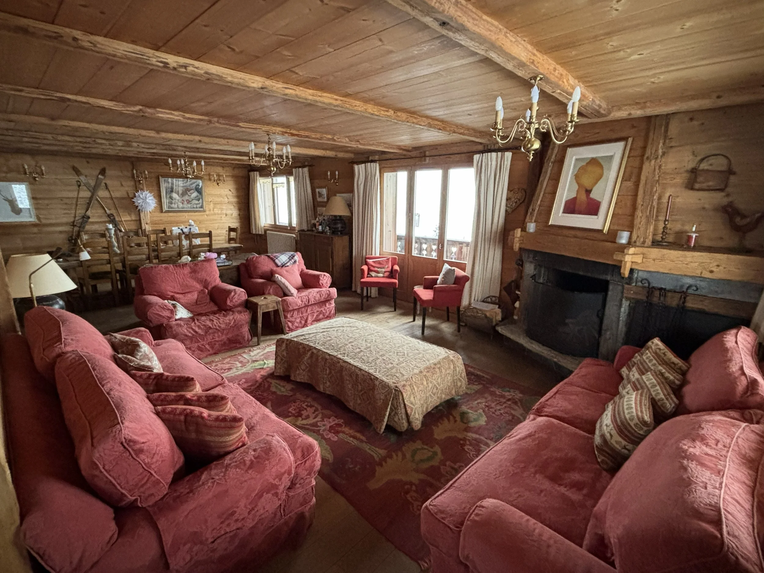 Living room with pink upholstered sofas and armchairs, a large area rug, a stone fireplace, wooden walls, ceiling, and furniture, with a chandelier hanging from the ceiling.