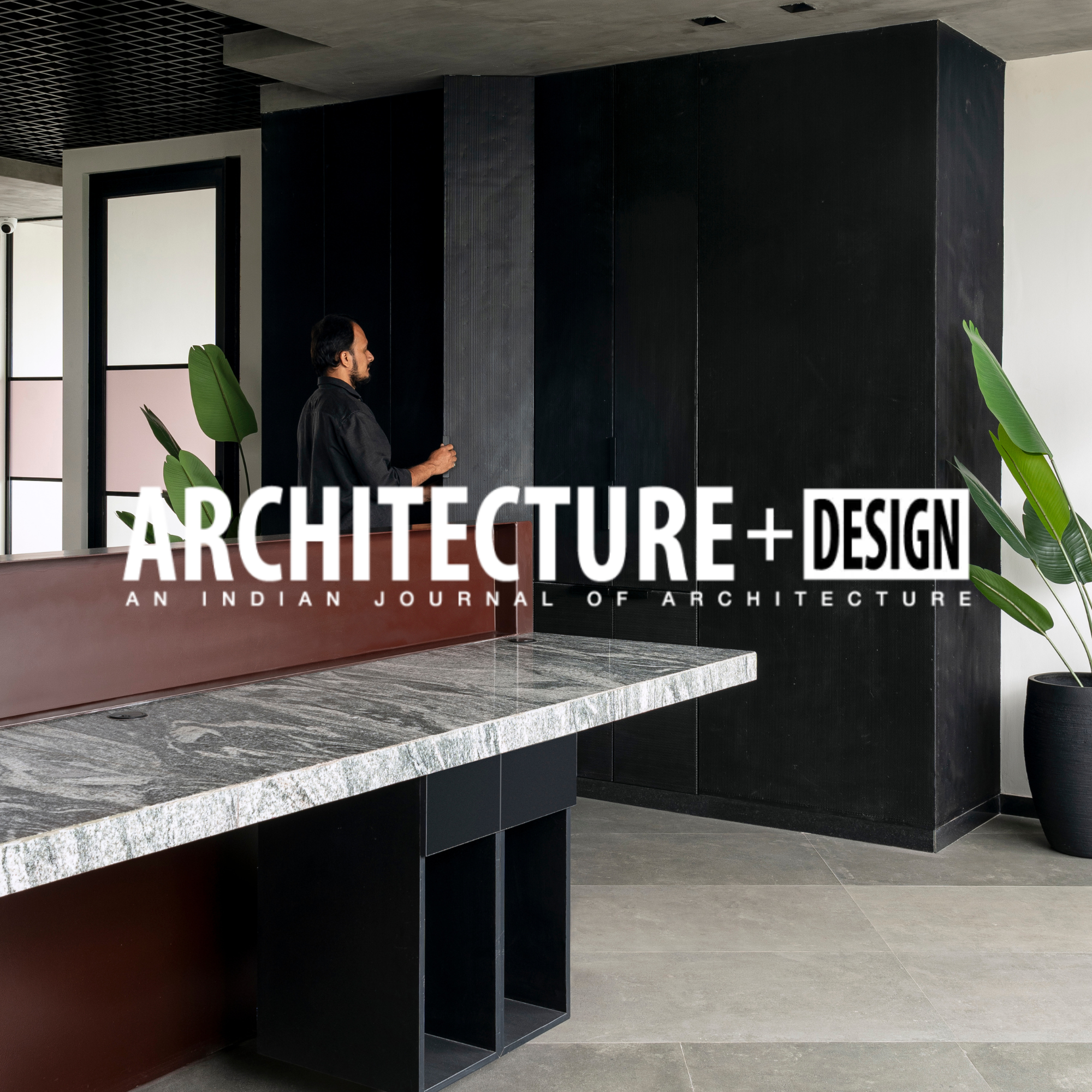Interior of a modern building with a man in black shirt near a black wall, large green potted plant, marble countertop, and large windows, with text overlay reading 'ARCHITECTURE + DESIGN: An Indian Journal of Architecture.'