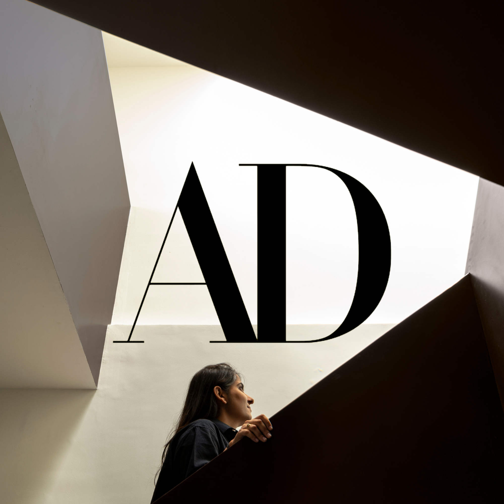 A woman with dark hair looking upward, holding onto a staircase railing, with a large black 'AD' on a white wall behind her, viewed from below.