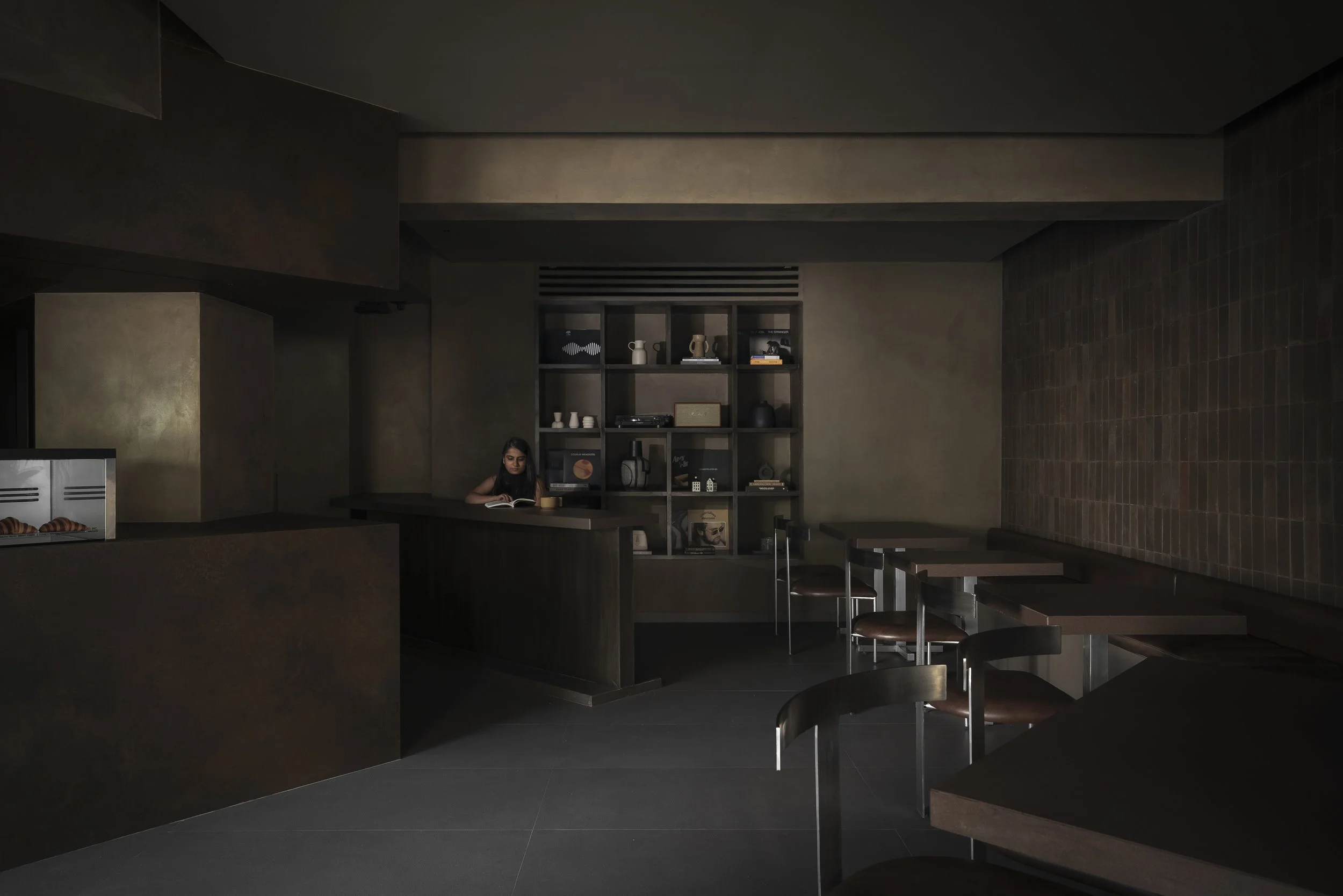 Interior of a dimly lit cafe or lounge with a woman sitting at a table reading a book, dark brown and gray color scheme, shelves with decorative items and records in the background, and several chairs and tables arranged throughout the space.