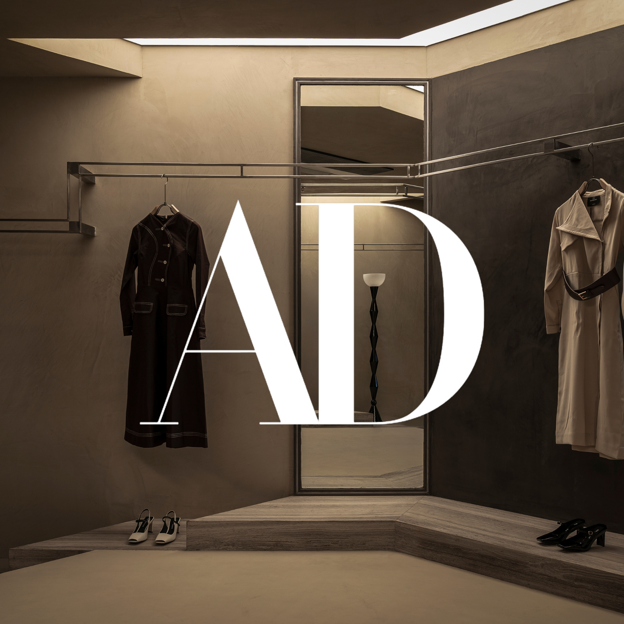 Clothing display with a black coat and white dress shoes on the left, and a beige trench coat and black heels on the right in a minimalist store with a large mirror and decorative floor lamp