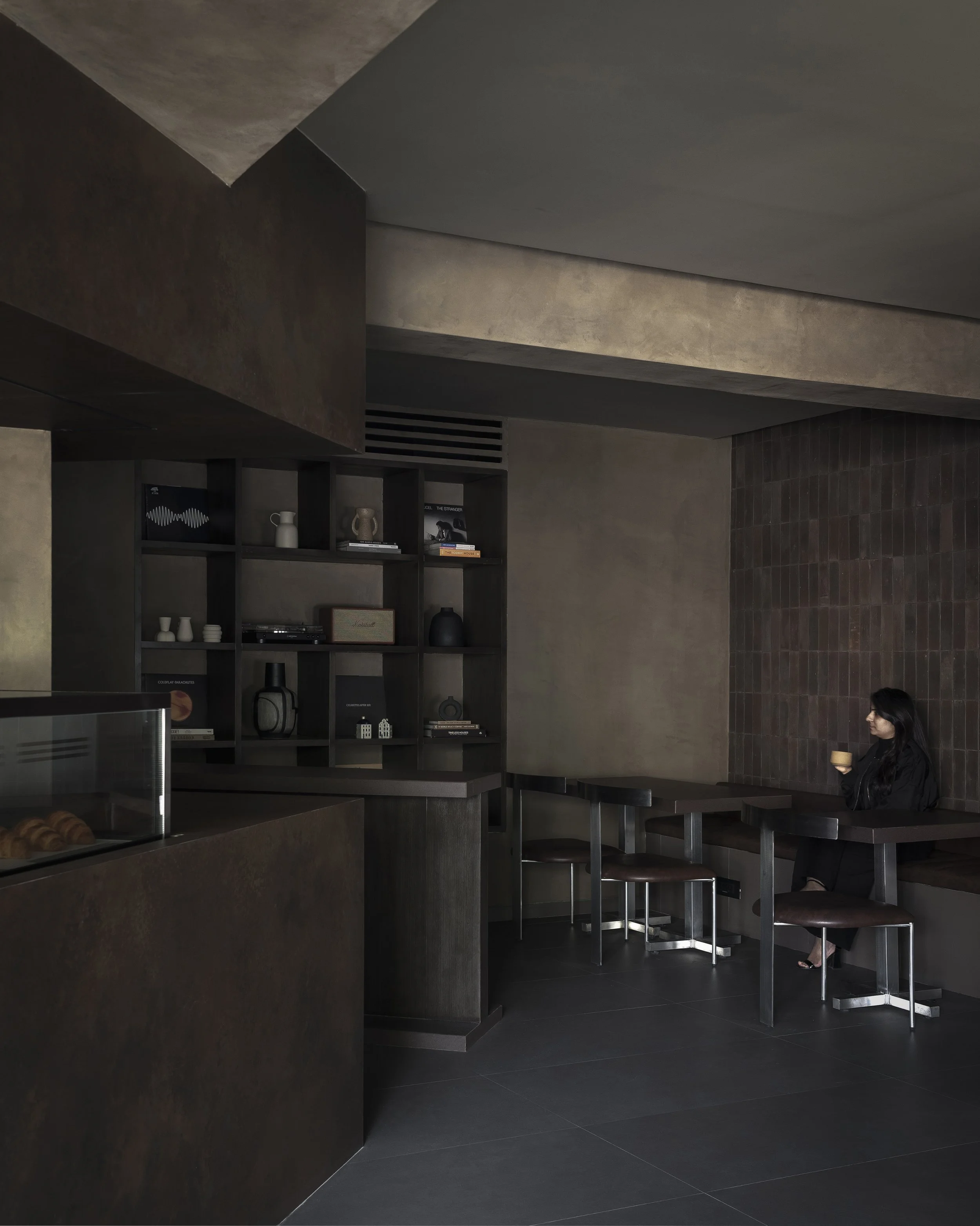 Modern interior coffee shop with large windows, sunlight casting shadows, and a woman sitting by the window reading.