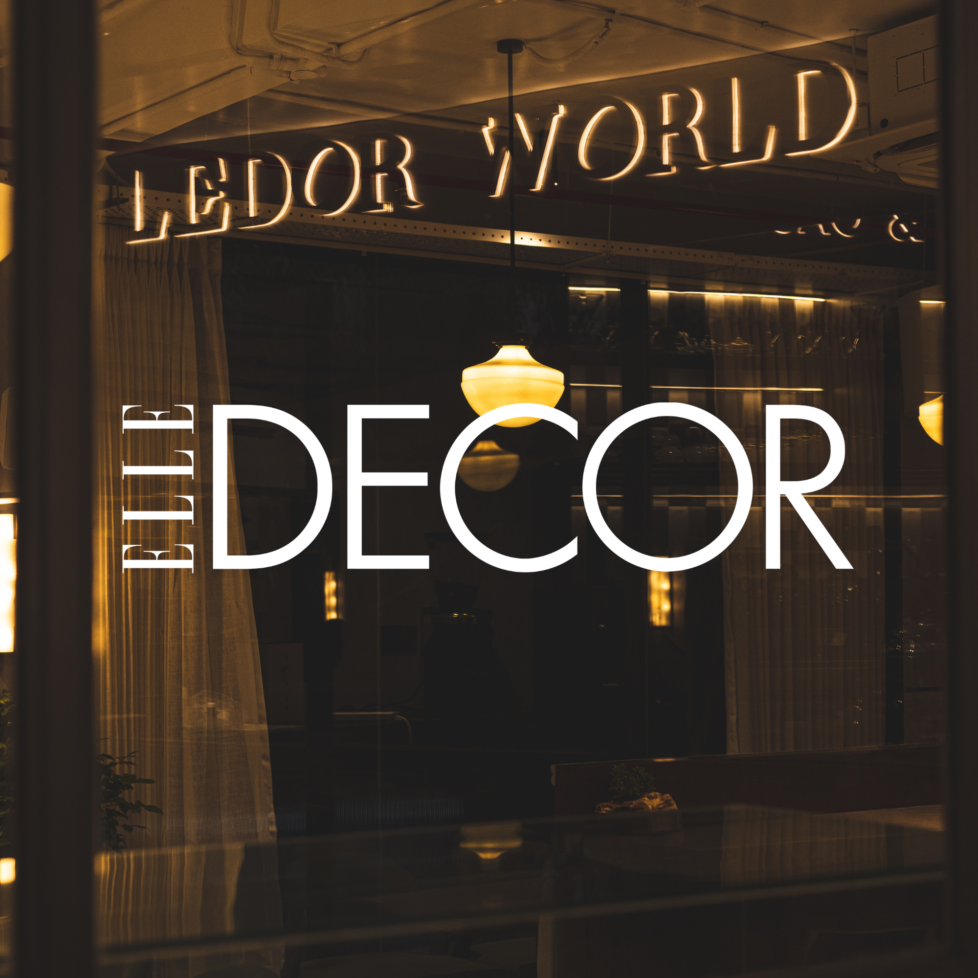 The window of an interior decoration store with illuminated signs reading 'E·DOR WORLD' and 'THE DECOR' visible through the glass, with warm lighting inside.