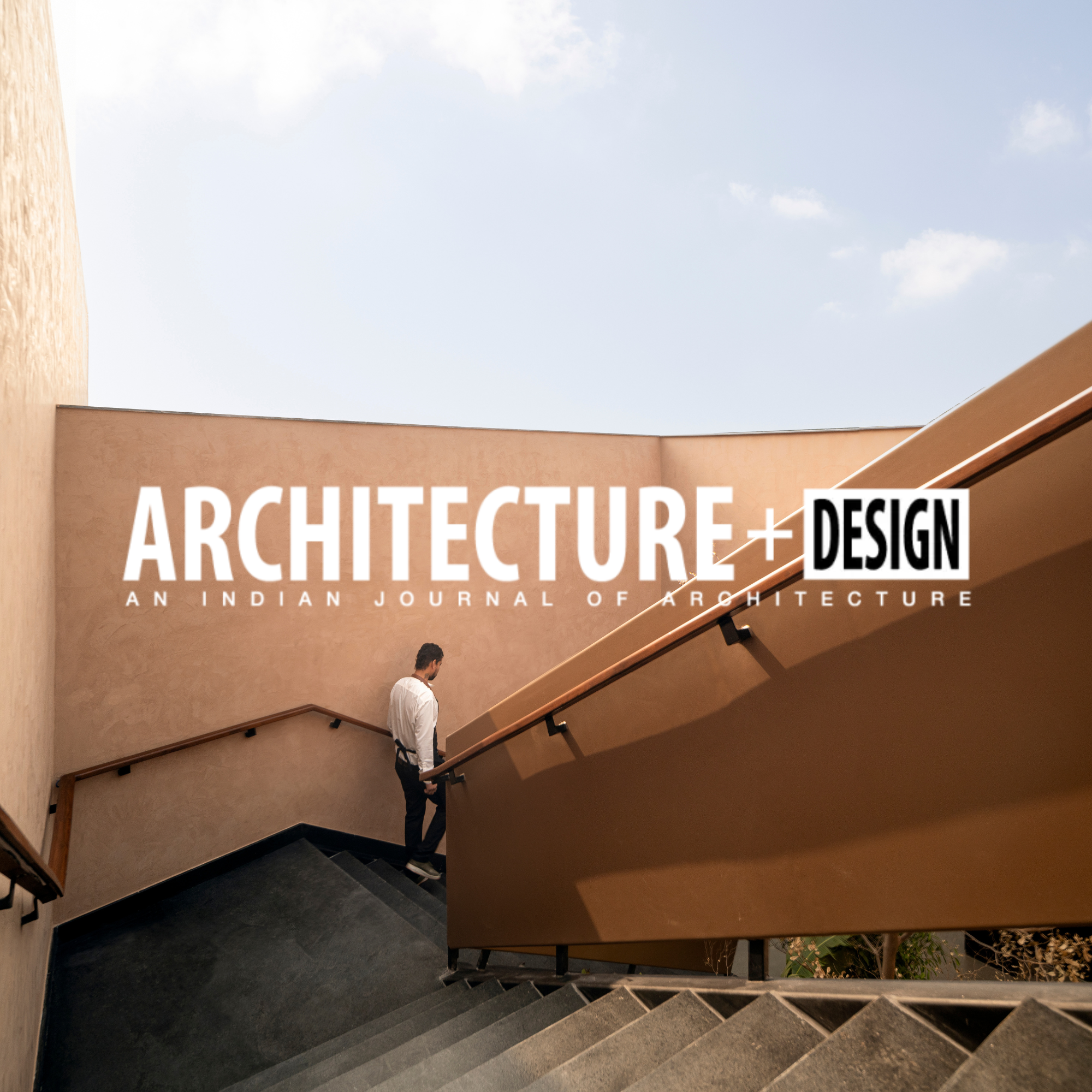 Man walking down a staircase outside with a modern, curved beige wall and a blue sky visible above, with the words 'Architecture + Design' and 'An Indian Journal of Architecture' overlaid.