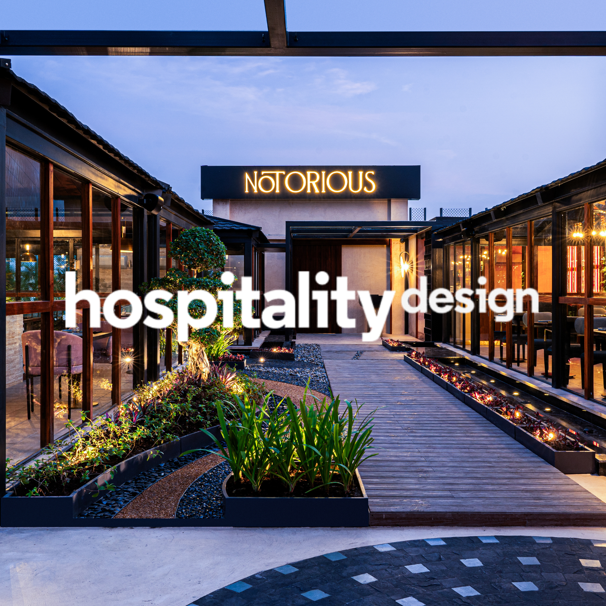 Exterior view of a modern restaurant or hospitality establishment with glass walls, wooden accents, and decorative plants, illuminated during dusk with a neon sign that reads 'NOTORIOUS' and overlay text that says 'hospitality design'.