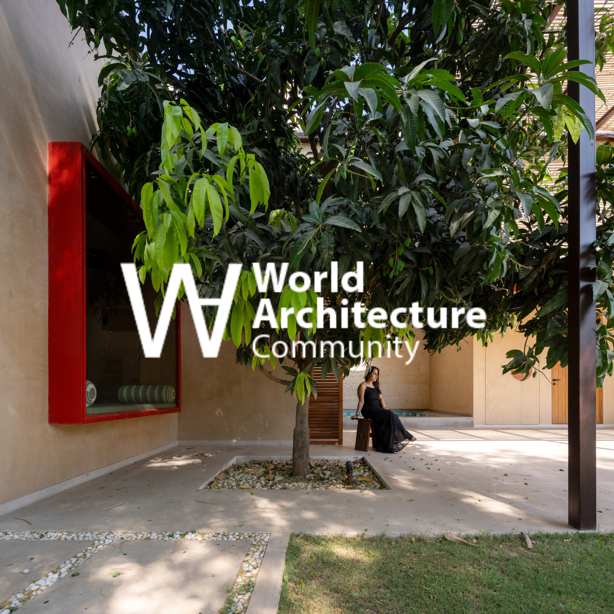 A woman in a black dress sitting on a bench under a large green tree in a modern courtyard with beige walls and a red window.