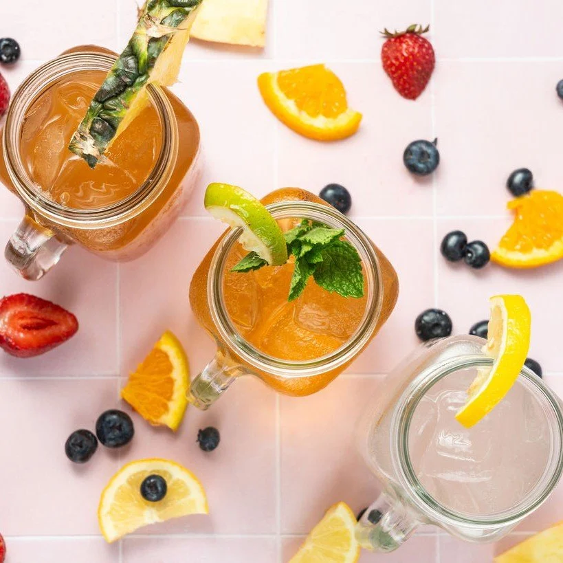 Three cocktails with fruit garnishes, surrounded by strawberries, oranges, and blueberries.