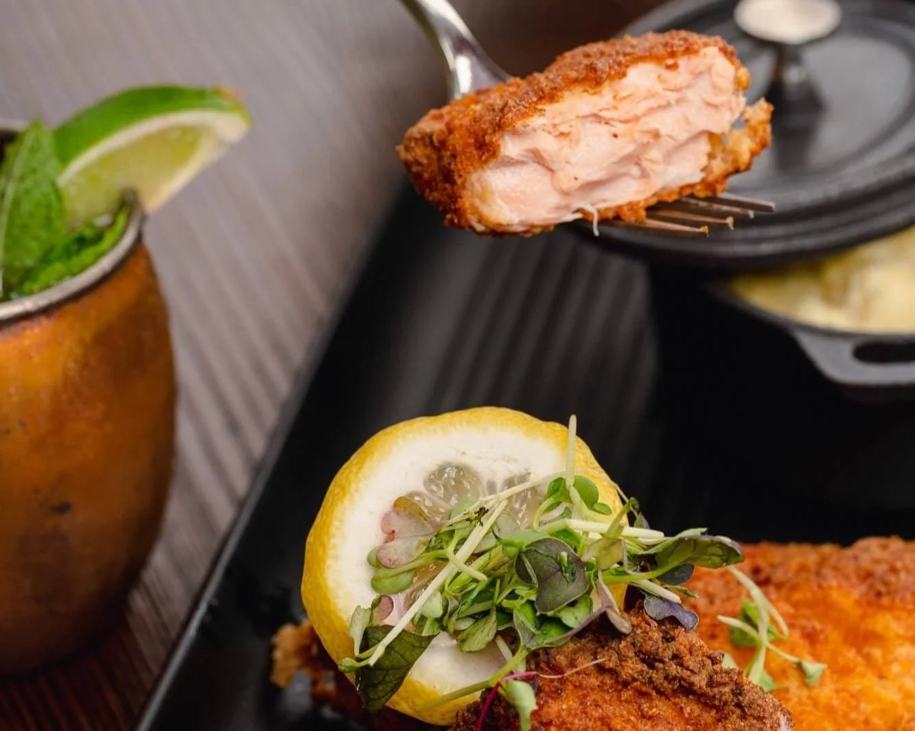 Close-up of a fork holding a piece of crispy fried fish with tender flaky inside, served with a lemon slice and fresh microgreens.