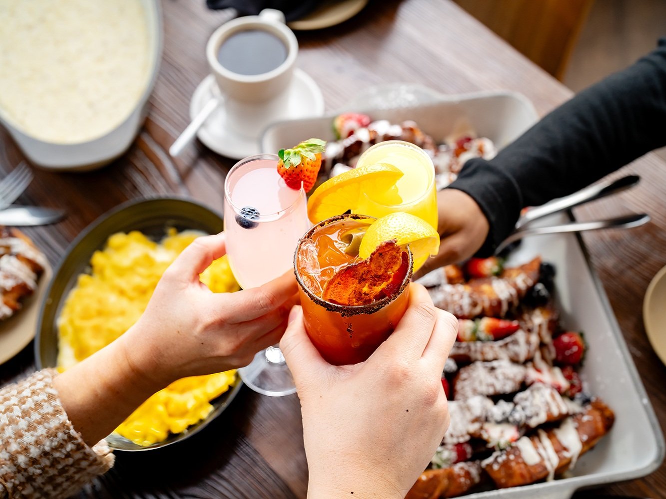 Hands clinking cocktails over a brunch table with eggs and pastries.