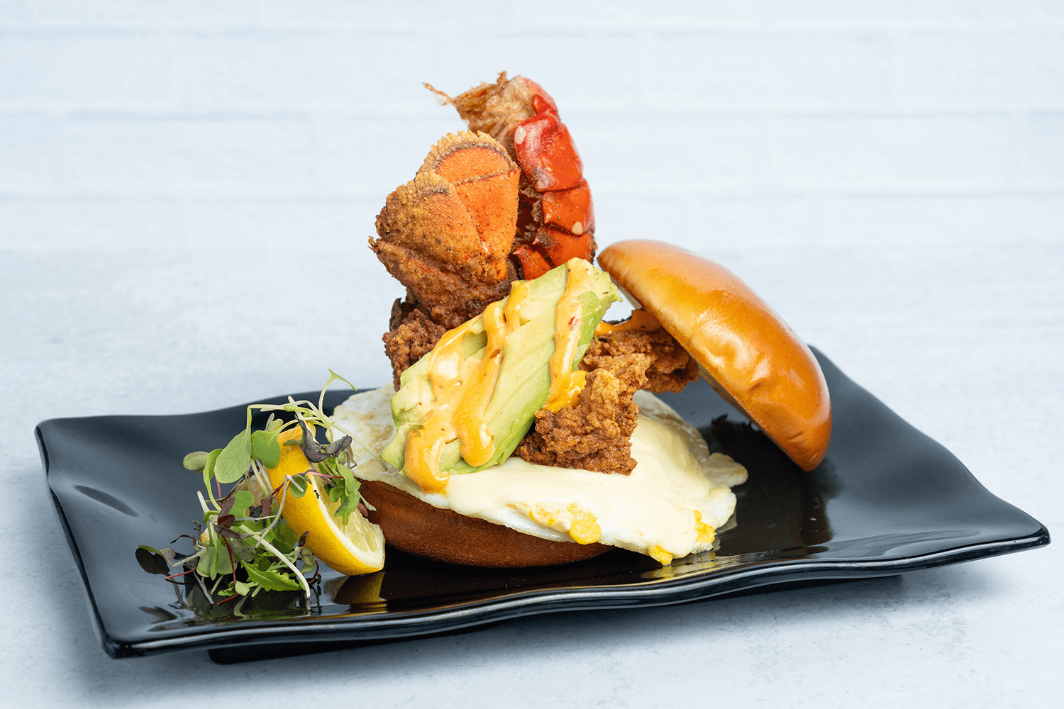 Lobster sandwich with avocado, sauce, and lemon garnish on a black plate.