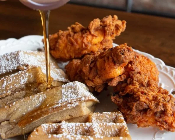 a person drizzling maple syrup on waffles and chicken.