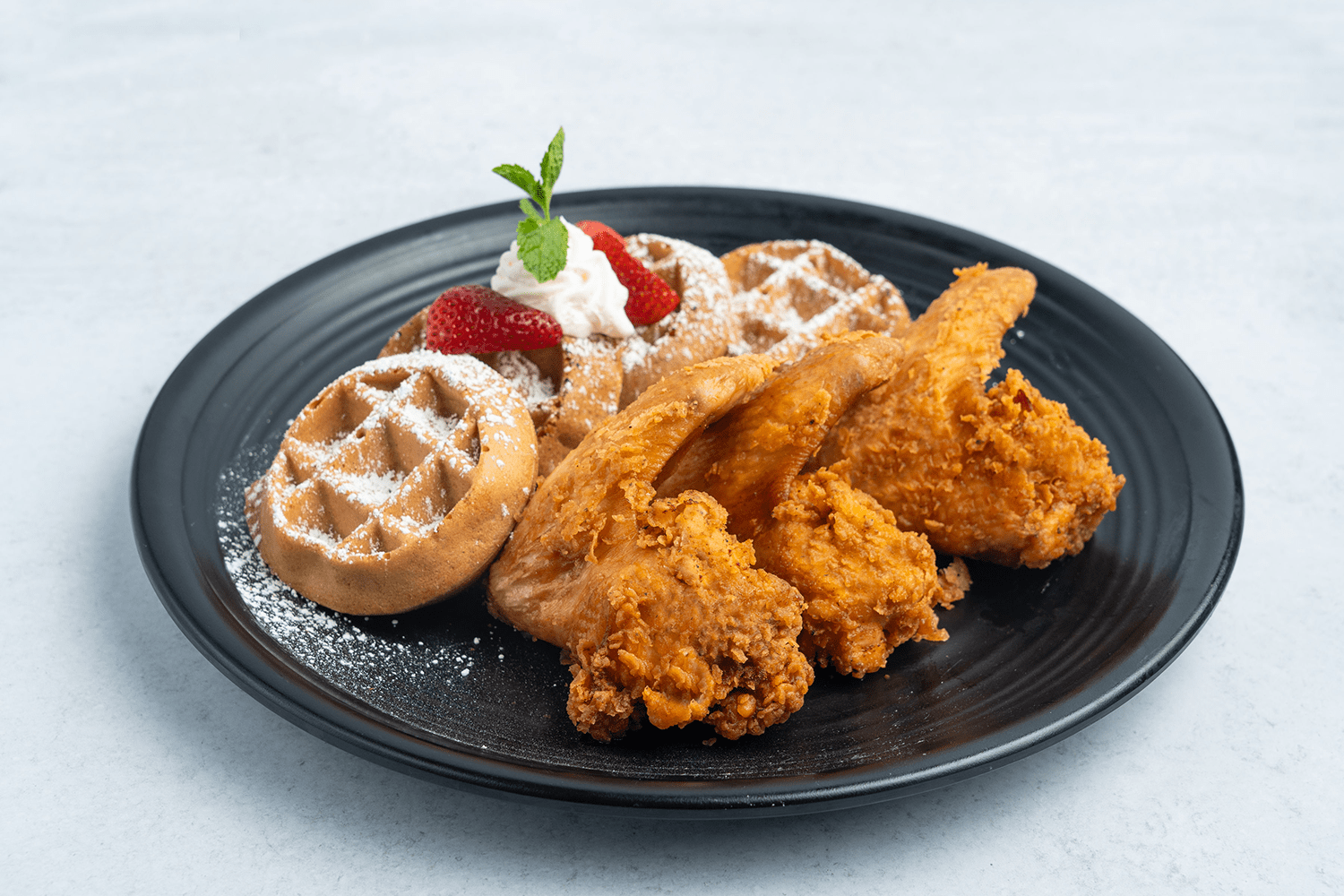 Classic Southern dish of crispy fried chicken and golden waffles topped with powdered sugar, fresh strawberries, and a sprig of mint, served on a black plate.