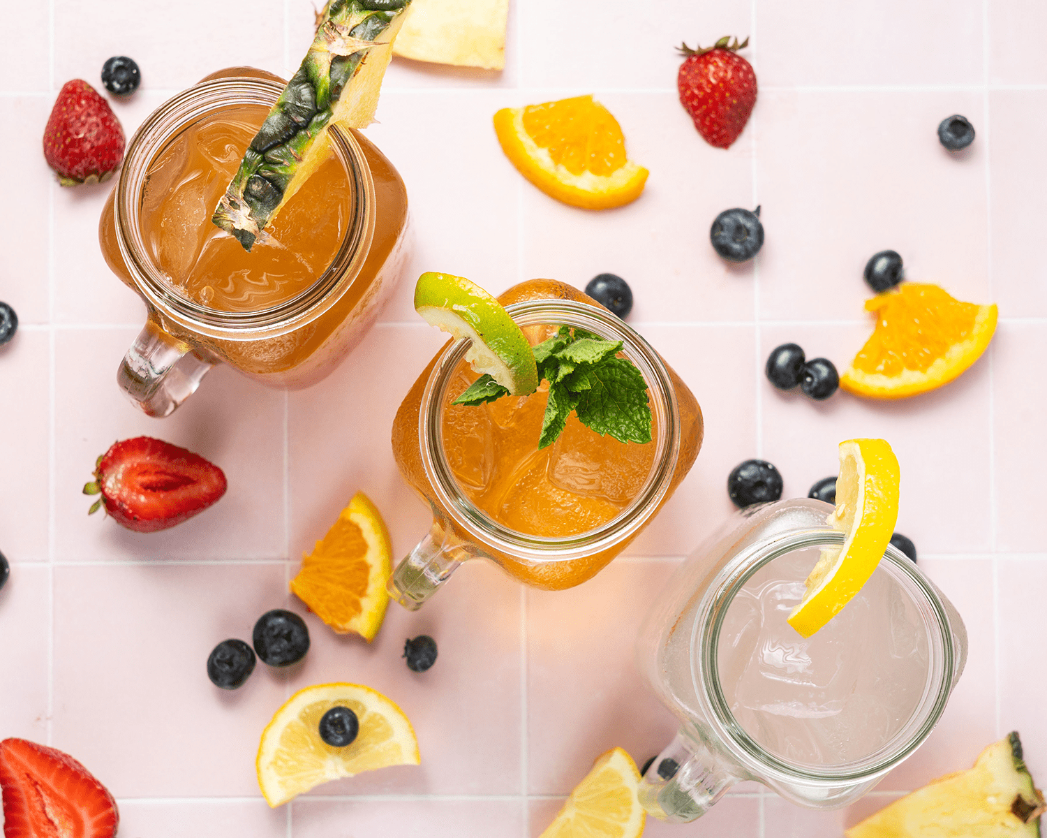 Three cocktails with fruit garnishes, surrounded by strawberries, oranges, and blueberries.