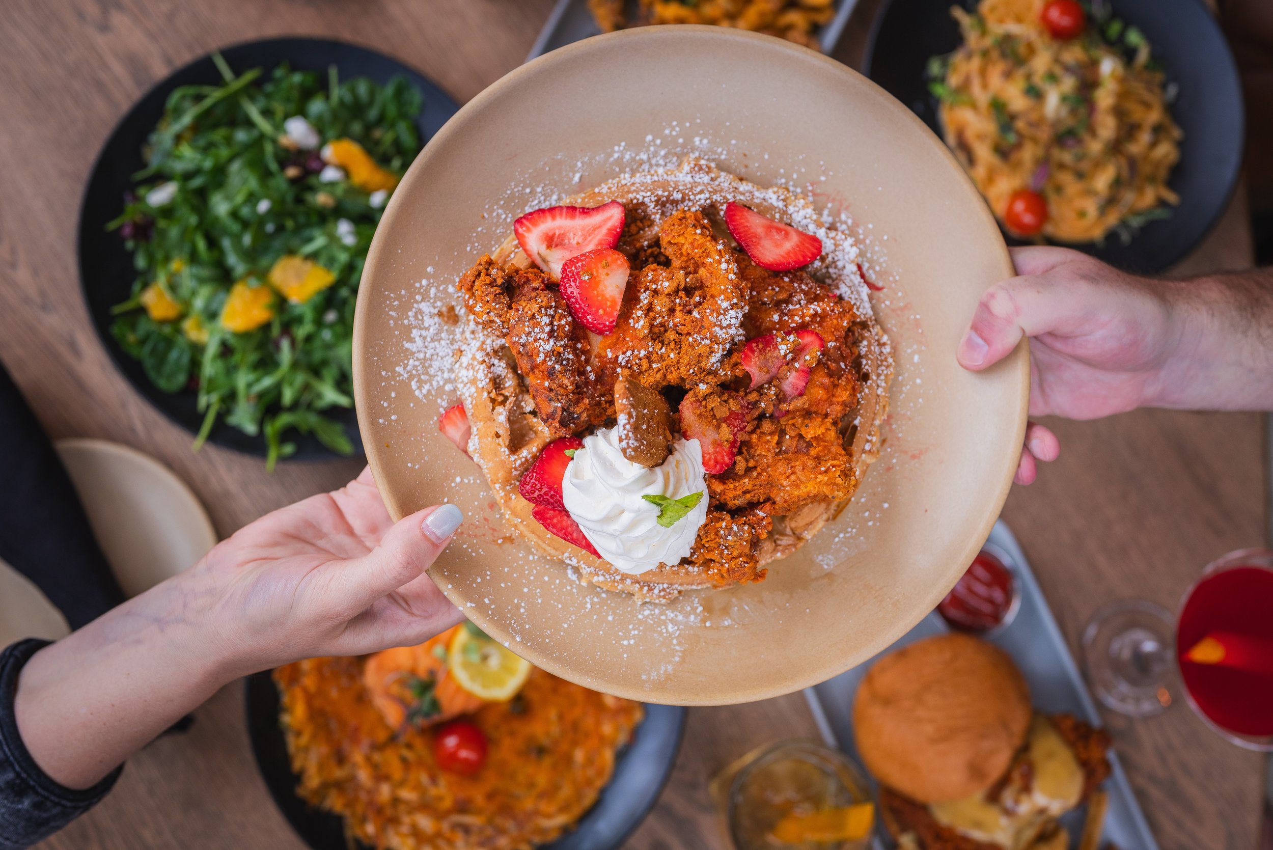 verhead view of multiple plated dishes on a wooden table, including fried chicken, greens, sausage patties, bacon, shrimp, and assorted sides.
