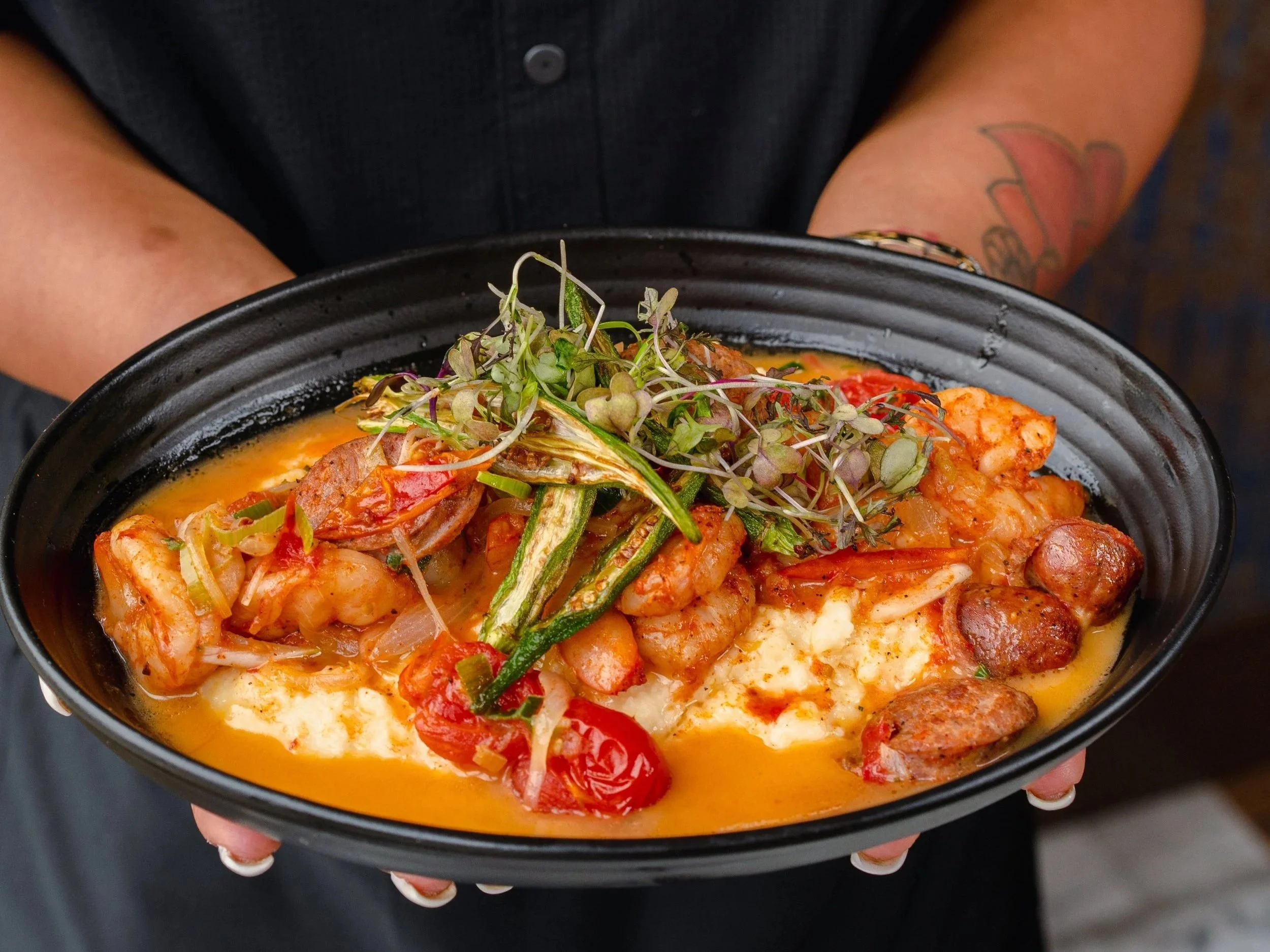 A person wearing a black button-up shirt with a yellow embroidered “Milk & Honey” logo holds a black bowl containing shrimp, sausage, cherry tomatoes, microgreens, and sauce over a creamy base.