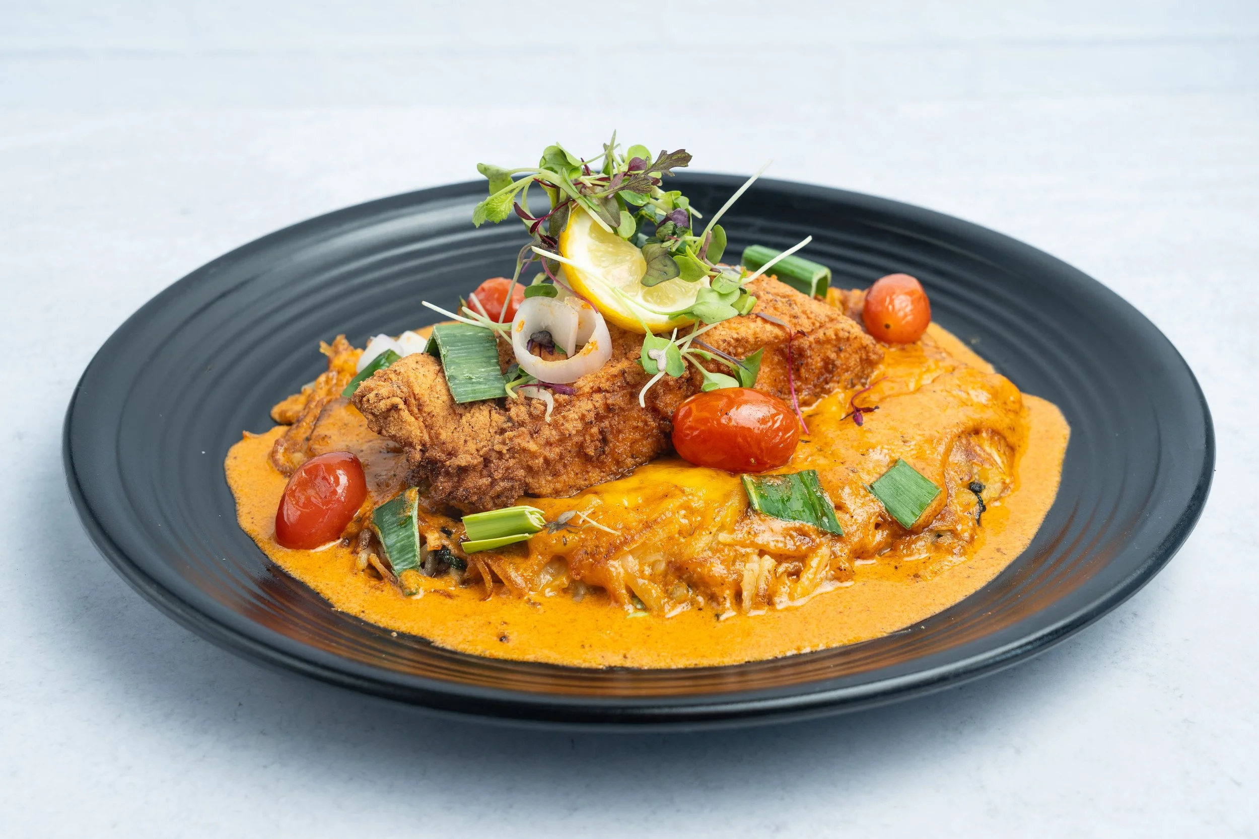 Crispy salmon fillet served over a hash-style base with cherry tomatoes, green onions, sliced leeks, and microgreens on a black plate with an orange-colored sauce.