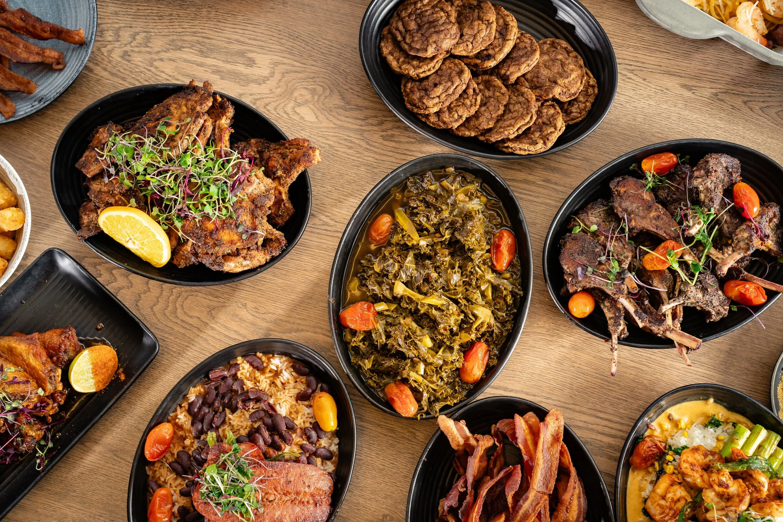 verhead view of multiple plated dishes on a wooden table, including fried chicken, greens, sausage patties, bacon, shrimp, and assorted sides.