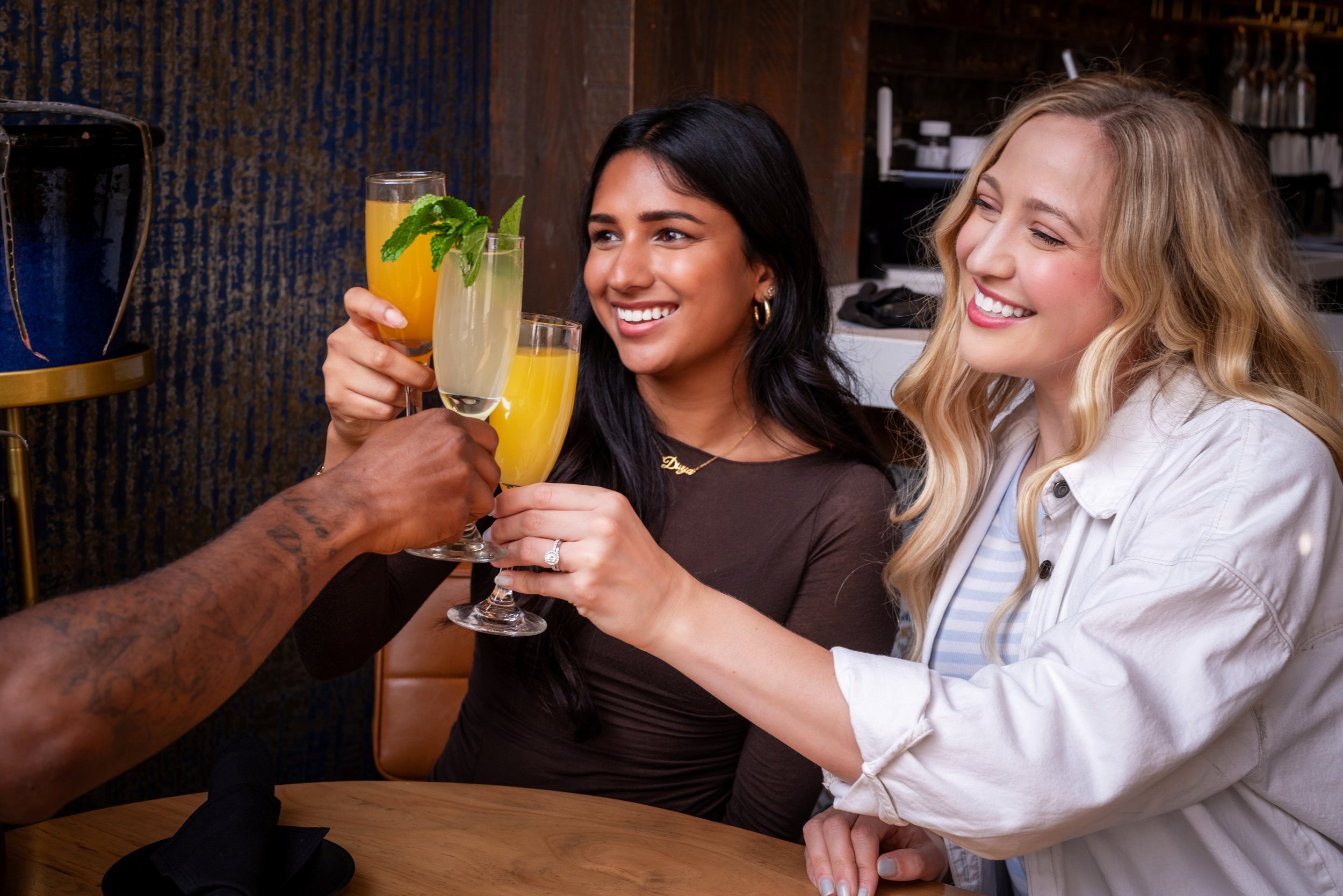 Four people raise champagne flutes filled with light-colored cocktails, one garnished with mint, in a dim restaurant bar setting, capturing a celebratory toast and smiling faces in front of a textured wall and barware.