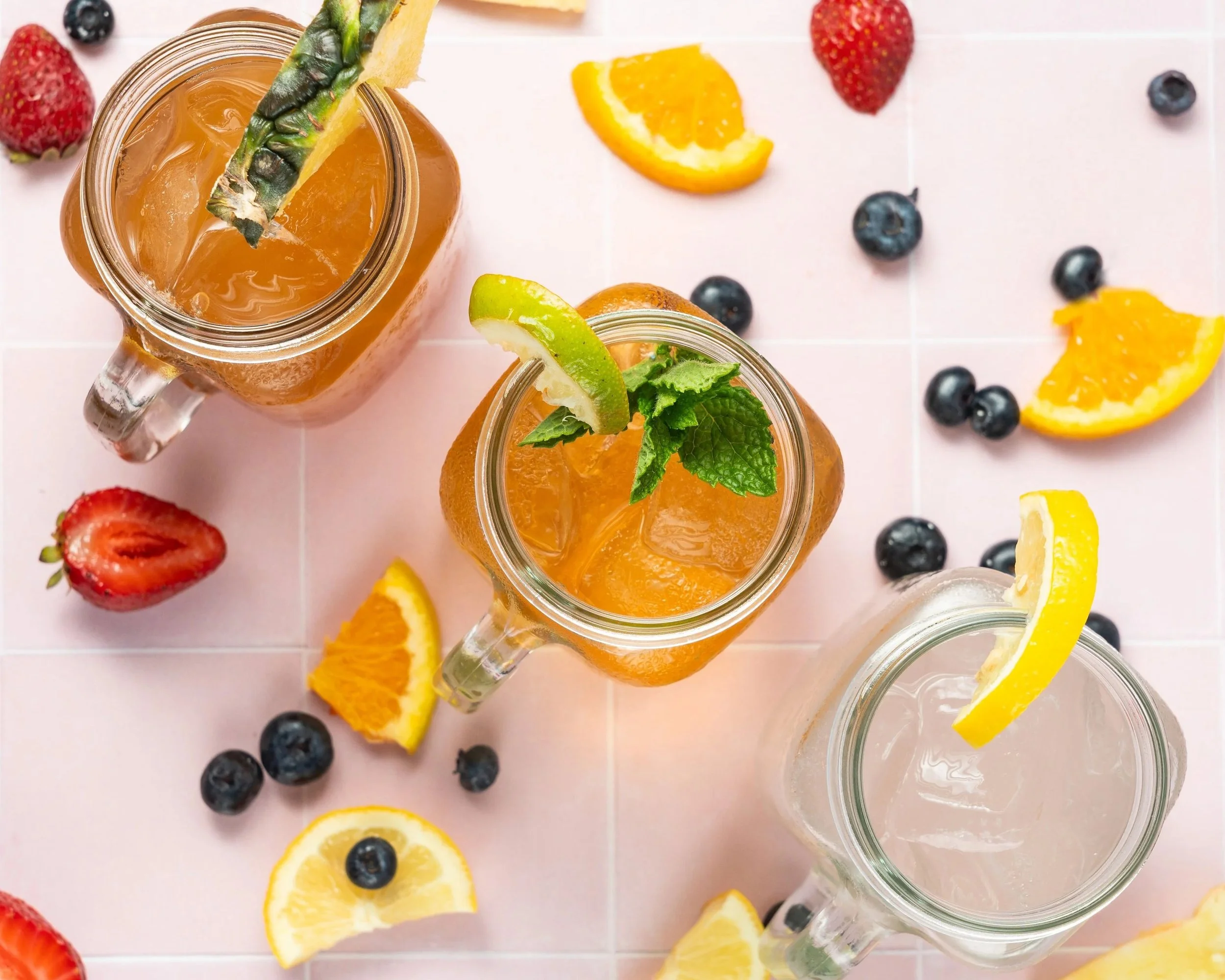 Top-down view of three glass jars containing iced beverages garnished with citrus slices and herbs, surrounded by scattered blueberries and sliced strawberries on a pink tiled surface.