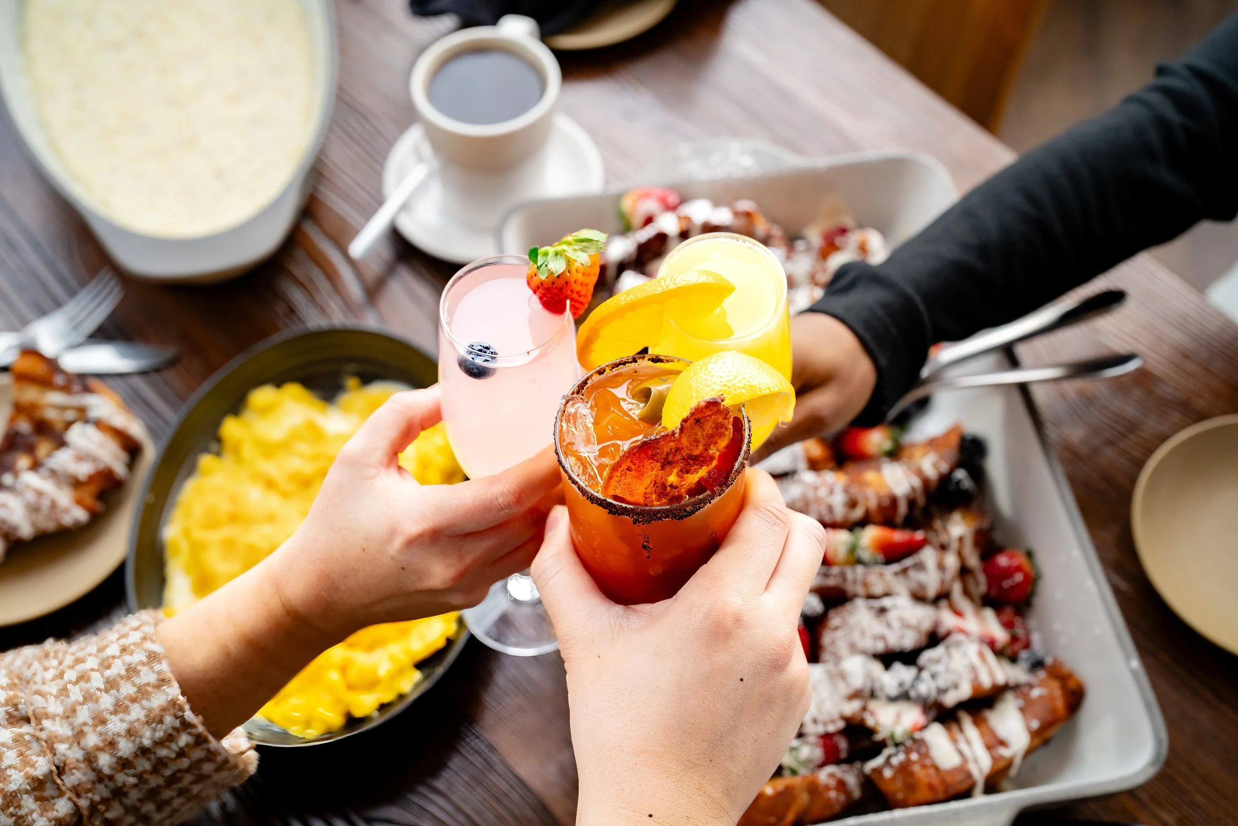 ree hands clink brunch cocktails over a table with scrambled eggs, pastries topped with powdered sugar and fruit, coffee, and breakfast plates.