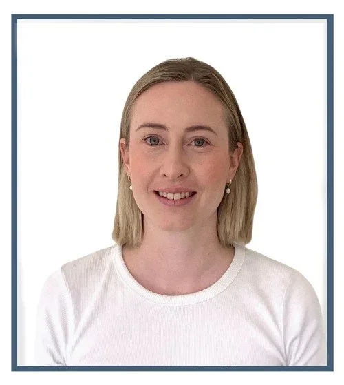 A young woman with blonde hair, wearing a white shirt and pearl earrings, smiling at the camera in front of a plain white wall.