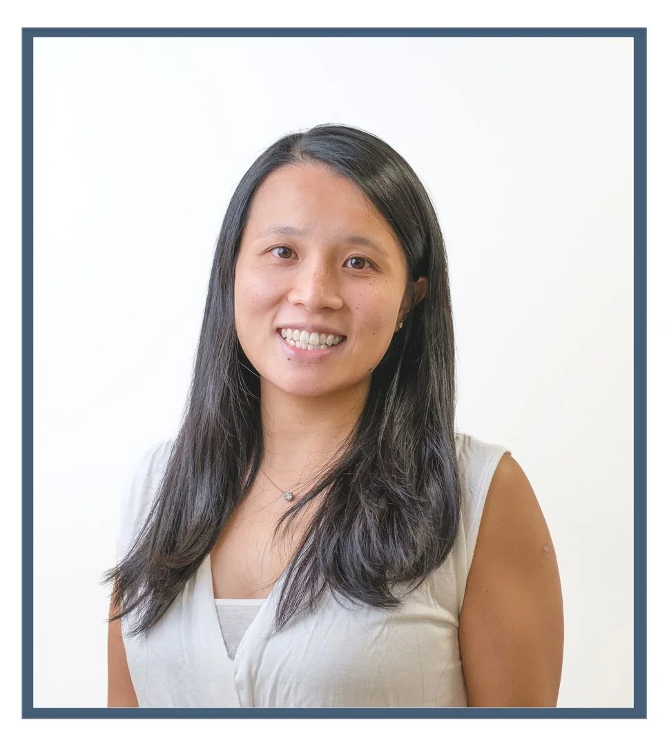 A woman with dark hair in a ponytail, wearing a navy polo shirt with a logo, standing against a plain white wall, smiling with hands on hips.