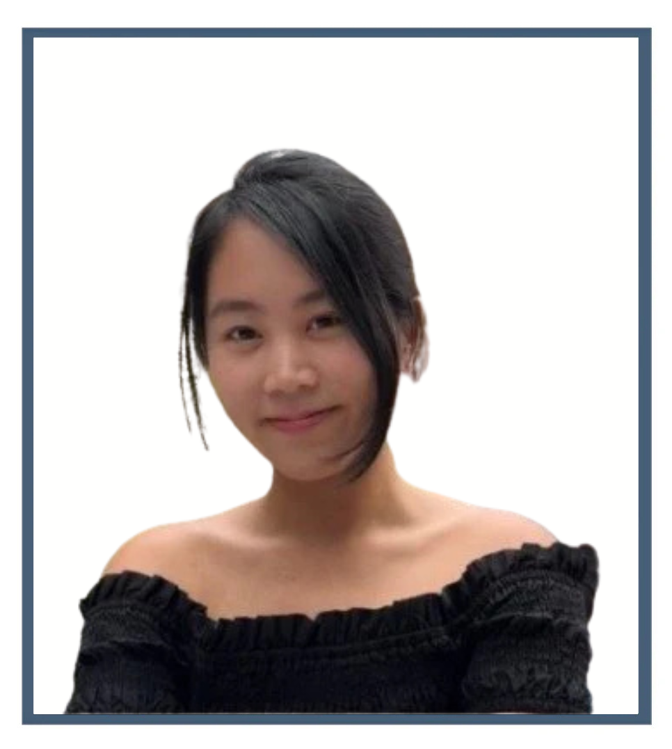 Young woman with short black hair wearing an off-the-shoulder black top, standing outdoors near a garden with umbrellas and someone in the background.