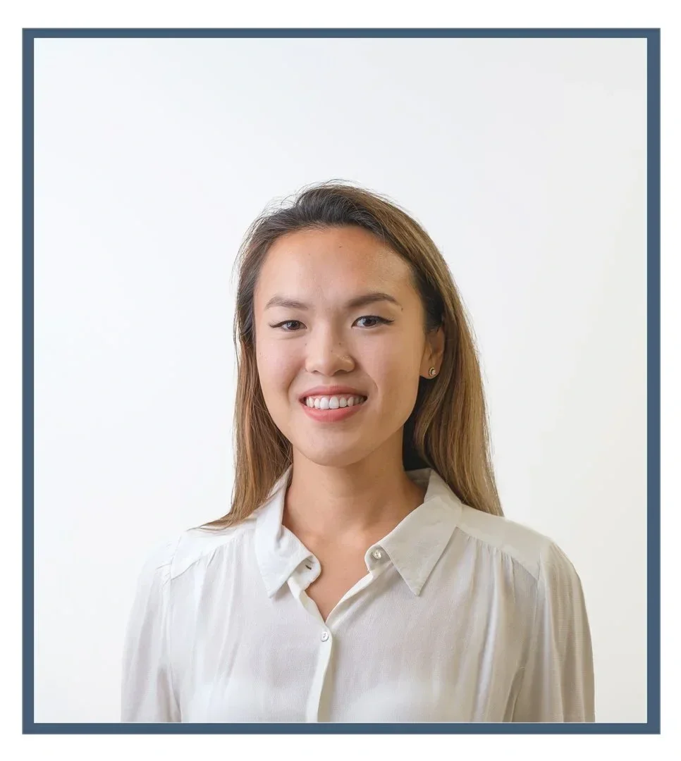 A young woman with dark hair tied up in a bun, smiling, wearing a navy blue polo shirt with a logo that reads "Joint Dynamics," standing against a wooden wall.