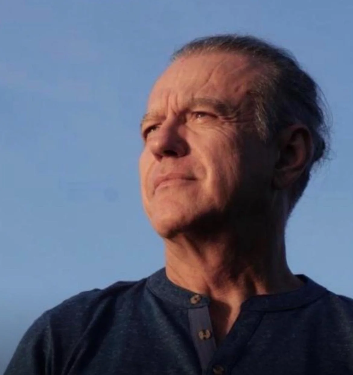 Un homme d'âge avancé regarde vers le ciel en plein jour, avec un air pensif, portant une chemise sombre.