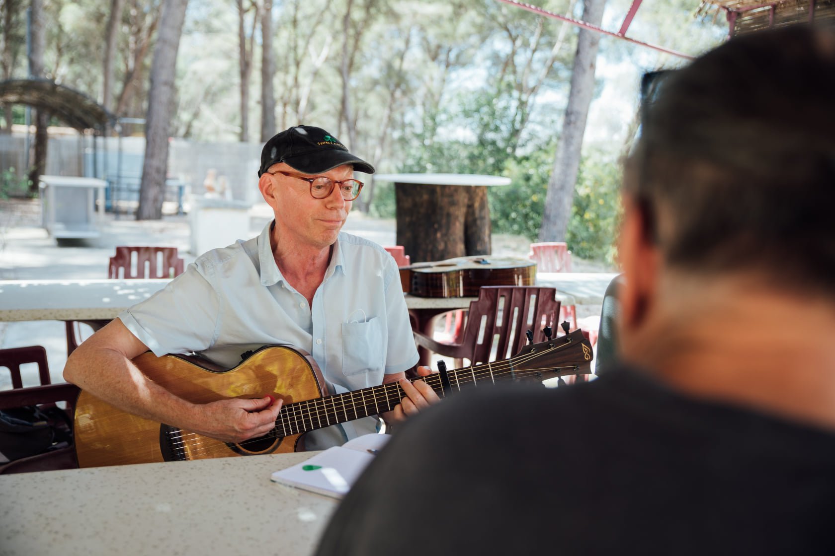 Un homme jouant de la guitare dans un lieu en plein air, assis à une table avec un groupe de personnes, sous un aménagement de bois et à proximité d'arbres.