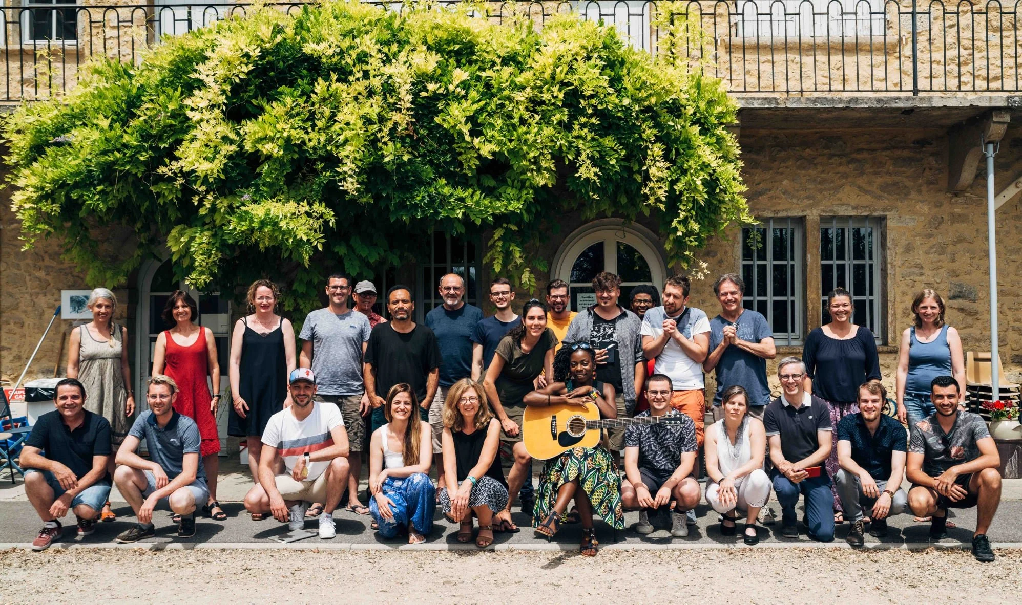Groupe de personnes posant devant une bâtisse en pierre avec une grande plante verte en arrière-plan, souriantes et détendues.