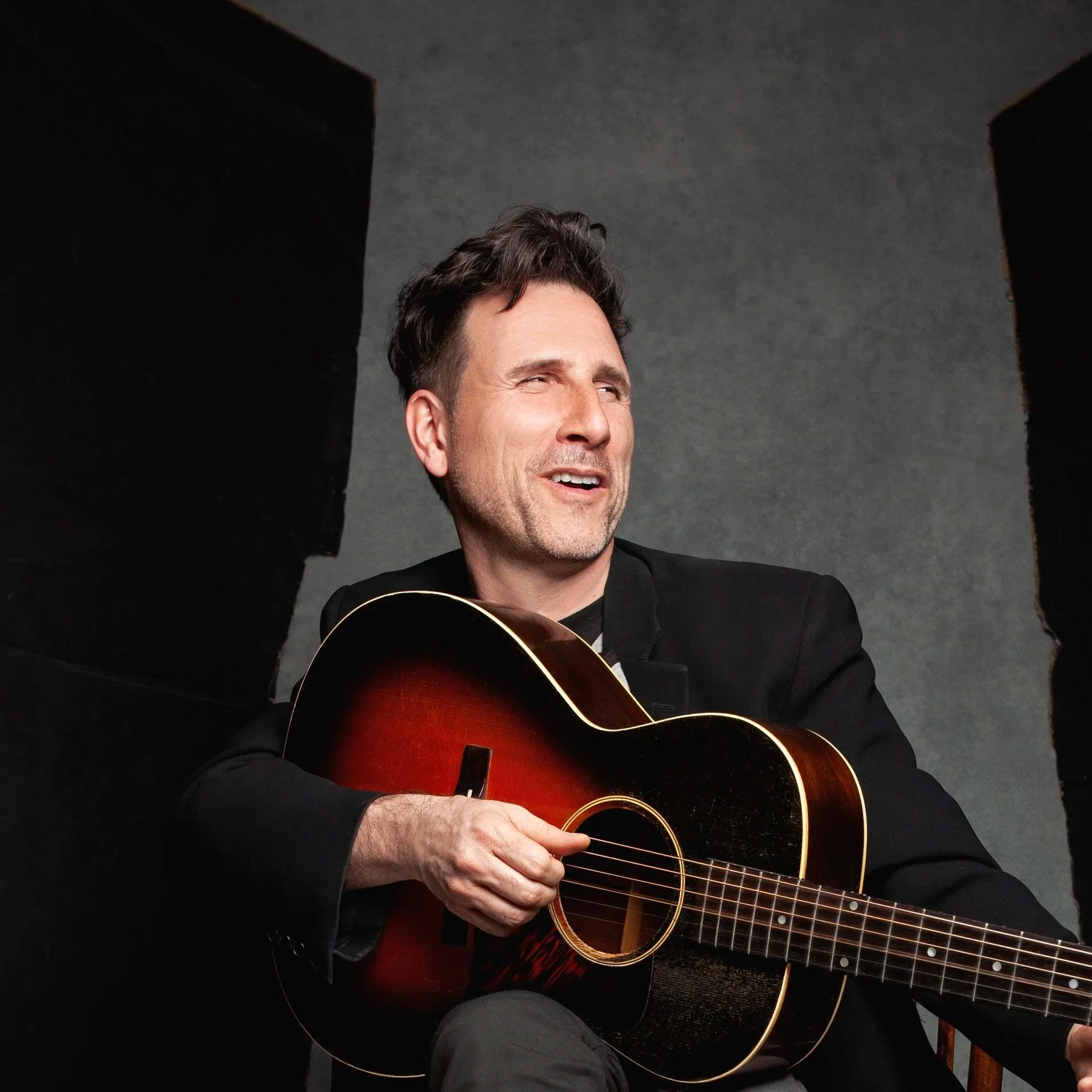 Homme souriant jouant de la guitare acoustique, assis devant un fond gris, entouré de grandes enceintes noires.