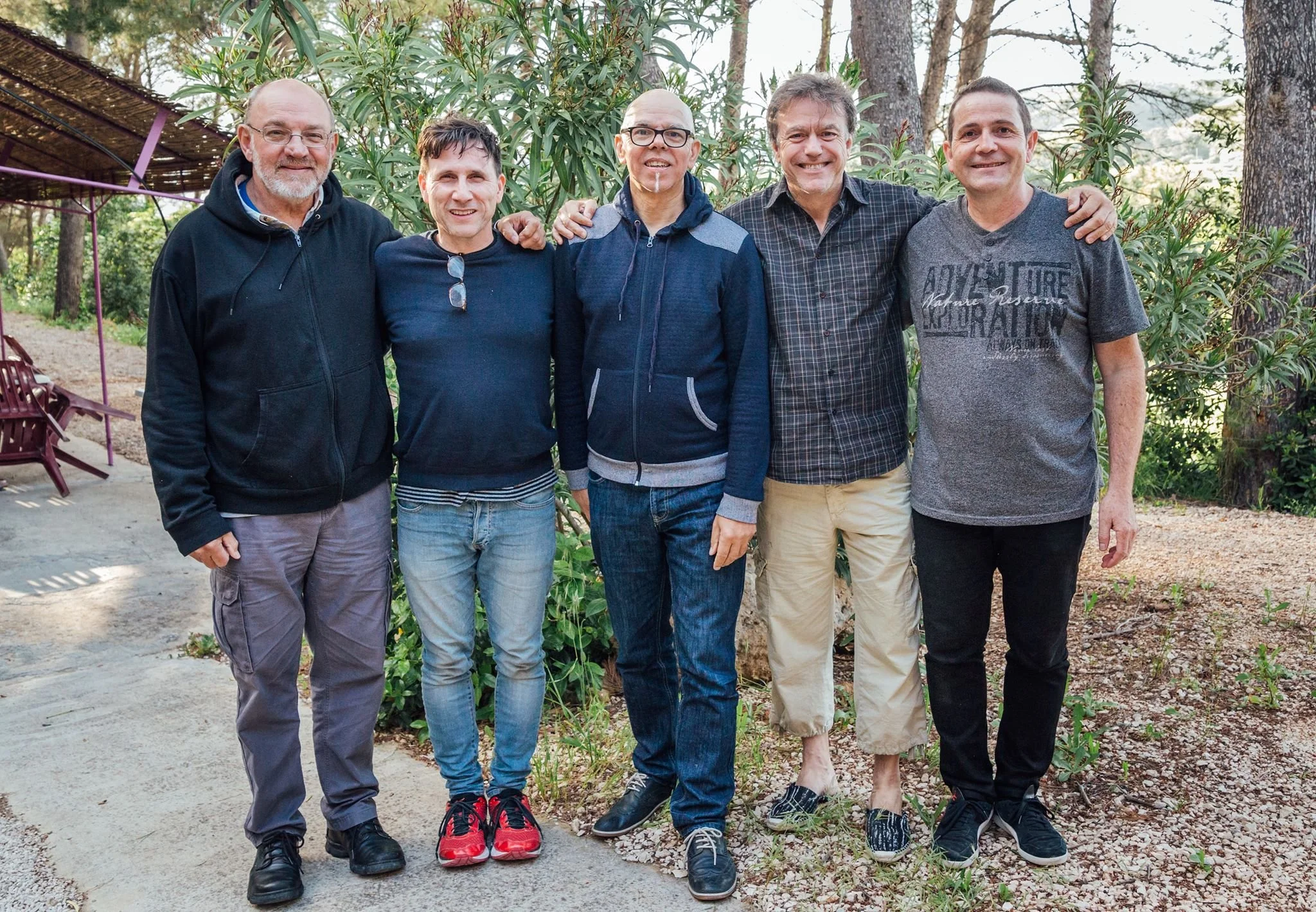 Groupe de six hommes souriants posant ensemble en plein air dans un jardin, avec des arbres et des meubles en bois en fond.