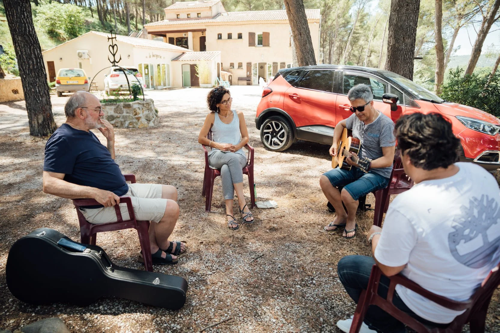 Un groupe de personnes joue de la musique et discute dans un espace extérieur ombragé avec des arbres et une voiture rouge en arrière-plan.