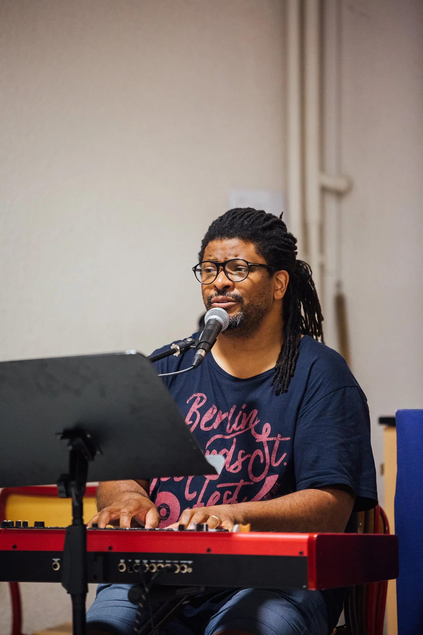 Homme jouant du clavier et chantant à la microphone, portant un t-shirt bleu avec du texte rose, dans un espace intérieur.