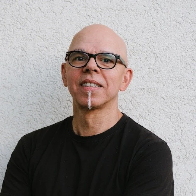Un homme portant des lunettes noires et un t-shirt noir sourit devant un mur blanc