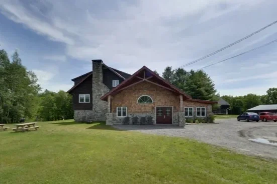 Maison en pierre avec toiture en pente, entourée de verdure et de voitures dans une allée, sous un ciel orageux.
