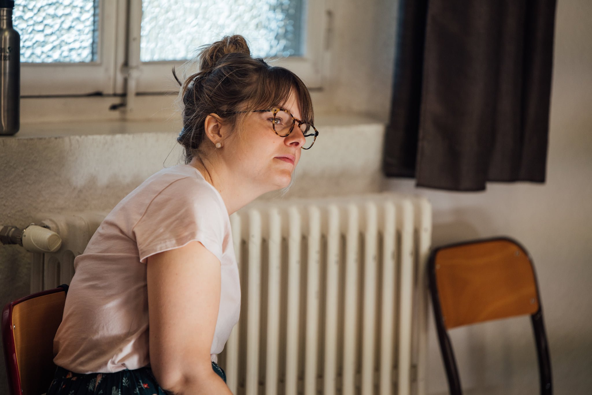 Une femme assise à côté d'une fenêtre, portant des lunettes, un t-shirt blanc et une jupe noire avec des fleurs, avec une expression pensive ou triste.