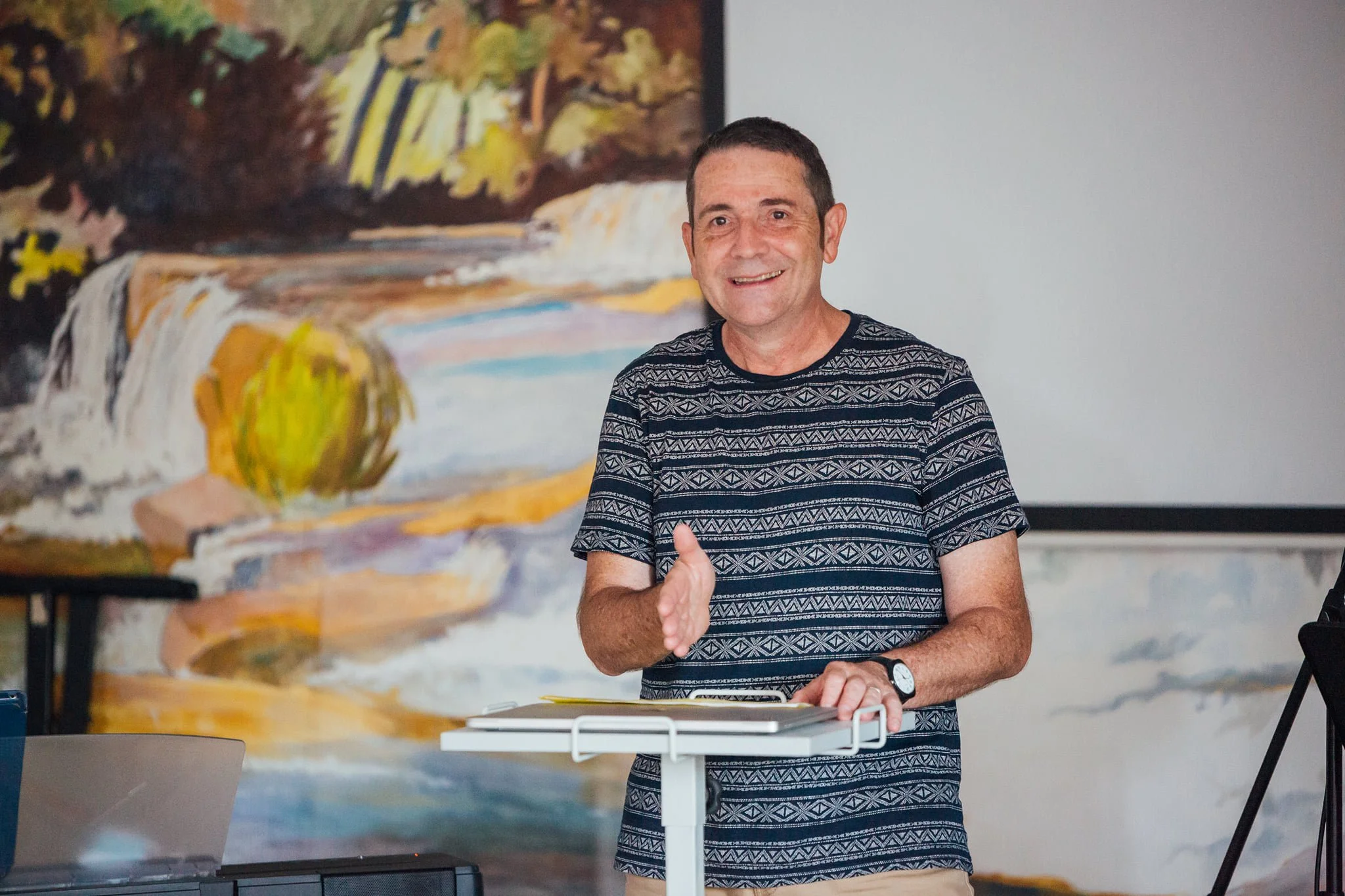 Homme souriant en train de parler, debout derrière un pupitre, en arrière-plan une peinture avec une cascade dans un paysage naturel.