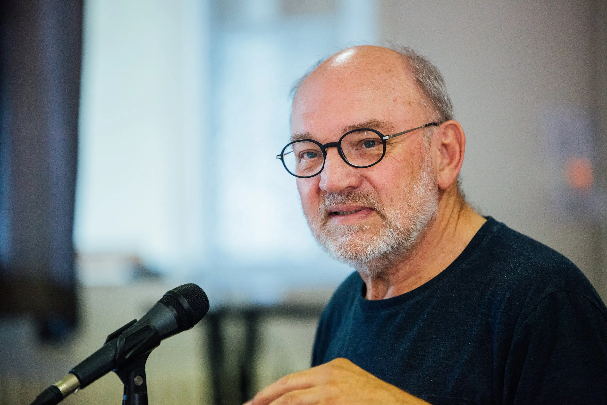 Un homme âgé avec des lunettes rondes, une barbe blanche, et portant un t-shirt noir, parle dans un microphone lors d'une réunion ou d'une présentation intérieure.