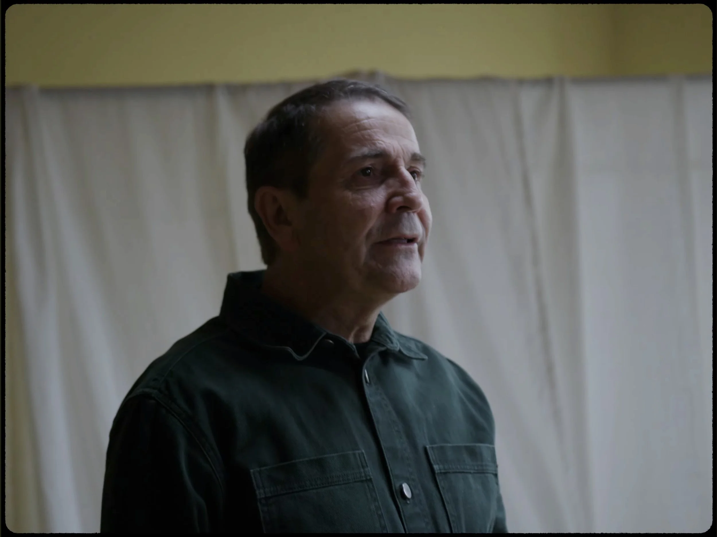 Un homme âgé avec des cheveux courts, portant une chemise noire, regarde vers la droite devant un rideau blanc, dans une pièce avec un mur beige.