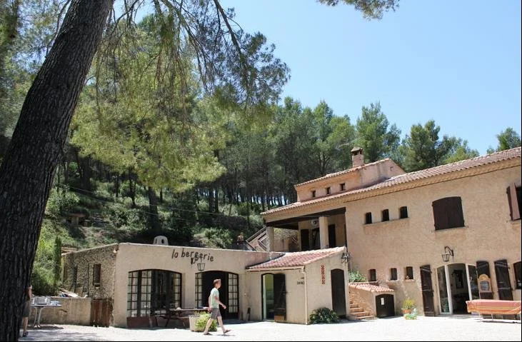 Un bâtiment de style provençal avec une terrasse extérieure en pierre, entouré d'arbres, et un homme marchant sur le chemin devant.