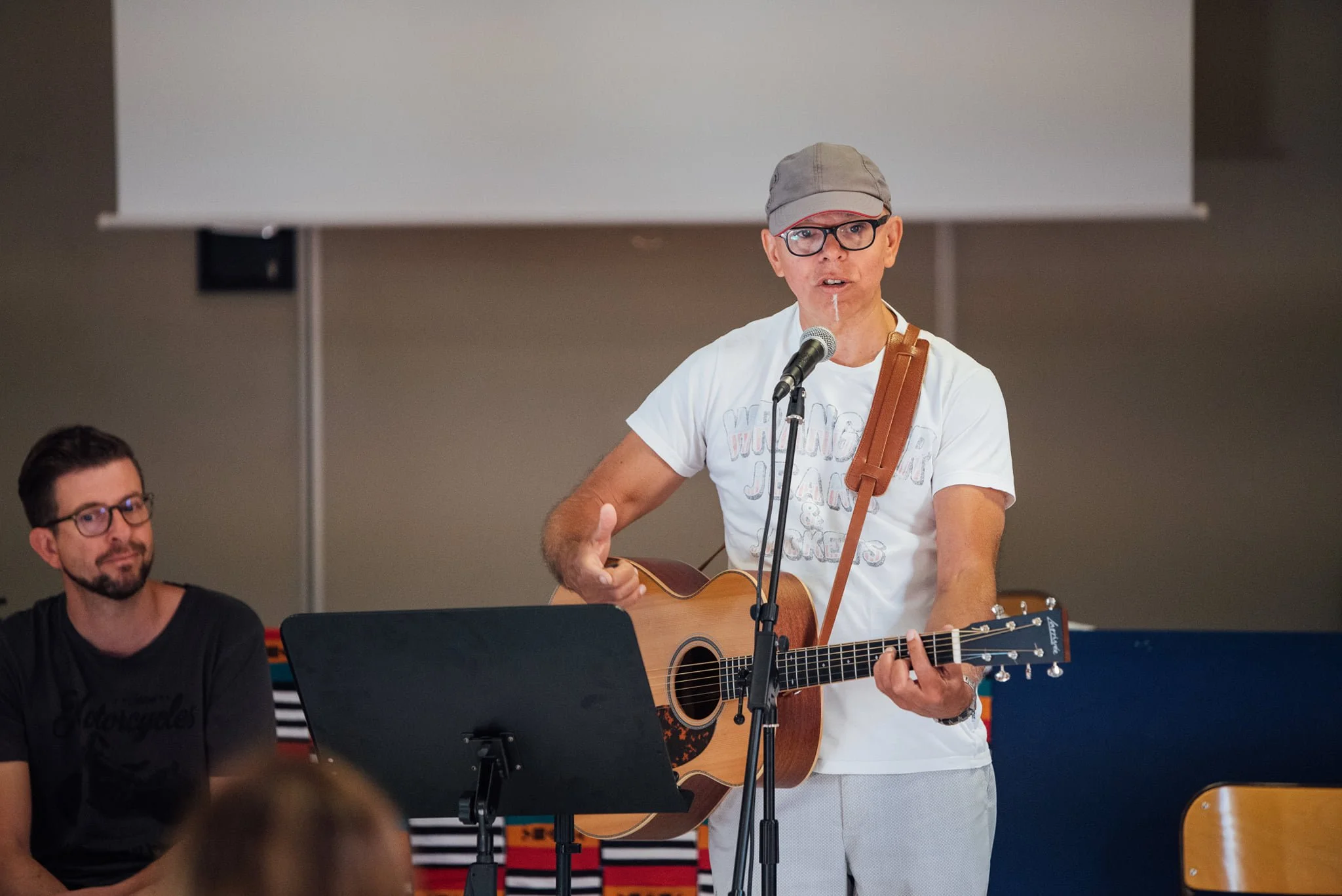 Un homme joue de la guitare acoustique et chante devant un microphone, un autre homme assis à côté le regarde, dans une salle de spectacle ou de répétition.