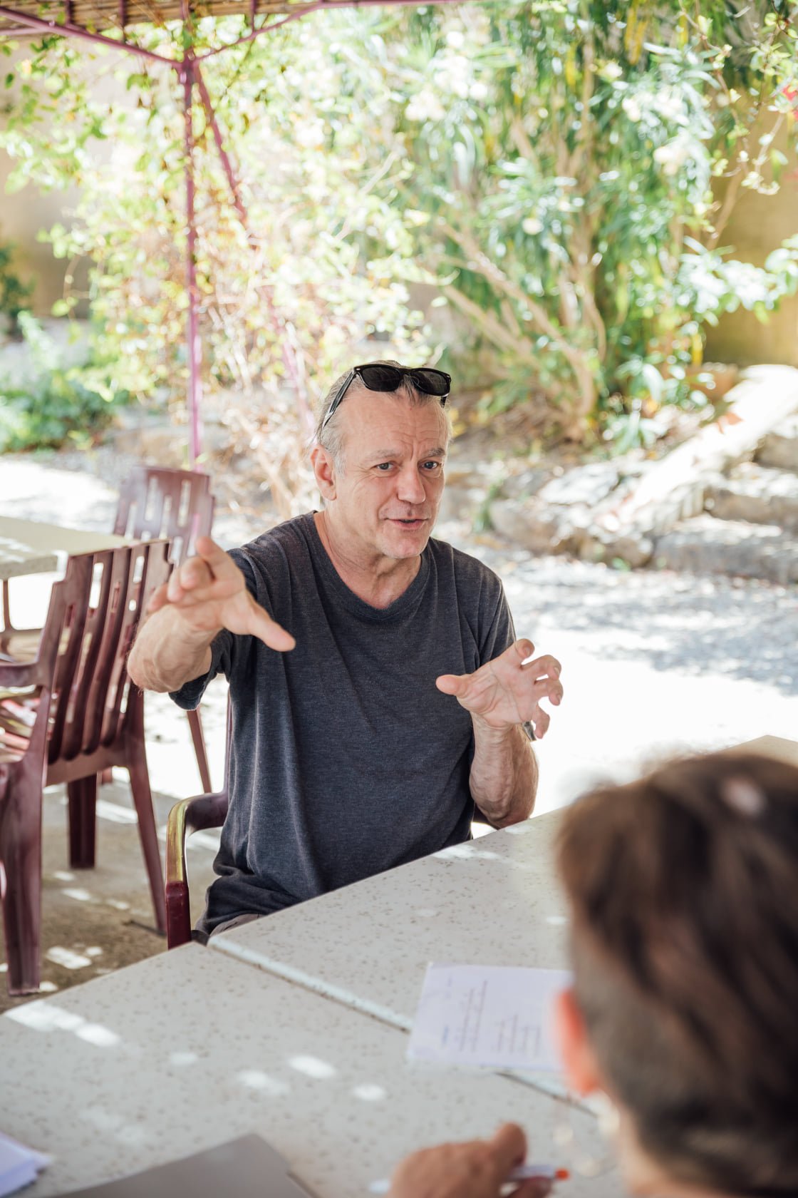 Un homme d'âge moyen discute avec une personne dont le dos est visible, assis à une table en extérieur sous un parasol, avec des plantes en arrière-plan.