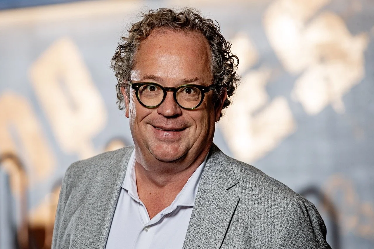 Man met krullend haar en bril, lachend, in een grijze jas en witte overhemd, met een achtergrond die lijkt op een muurschildering of kunstwerk.
