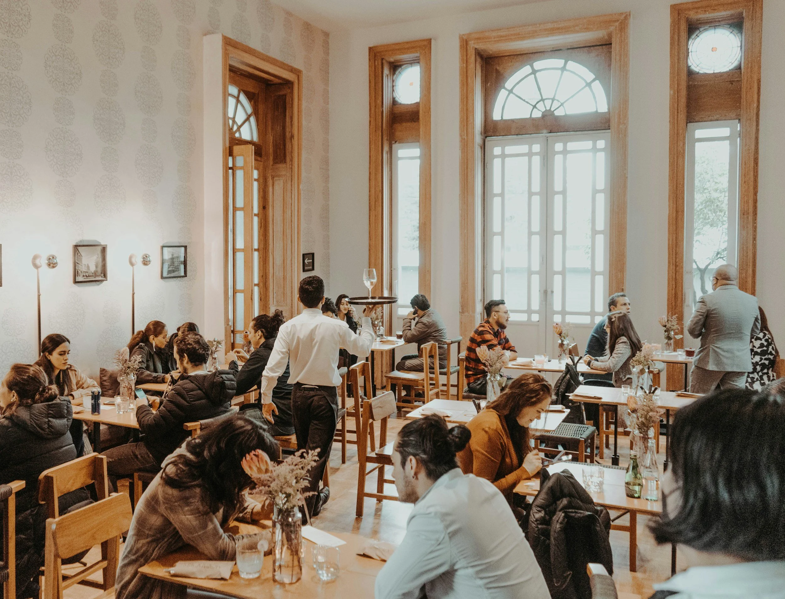 Interieur van een restaurant of cafe met hoge ramen, mensen die aan tafels zitten en een ober die serveert.