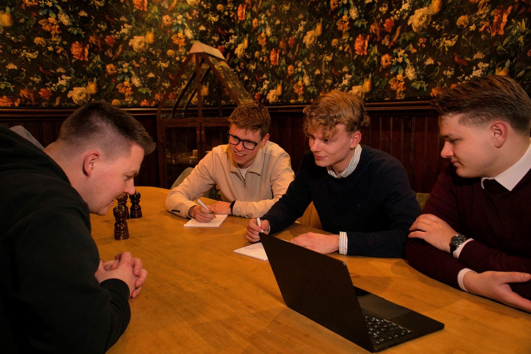 Vier jonge mannen zitten aan een houten tafel in een gezellig café of restaurant, met een bloemmotief muur op de achtergrond. Ze lijken een bordspel te spelen en zijn in gesprek of lachen. Een van hen heeft een laptop open voor zich. Ze dragen casual tot semi-formele kleding.