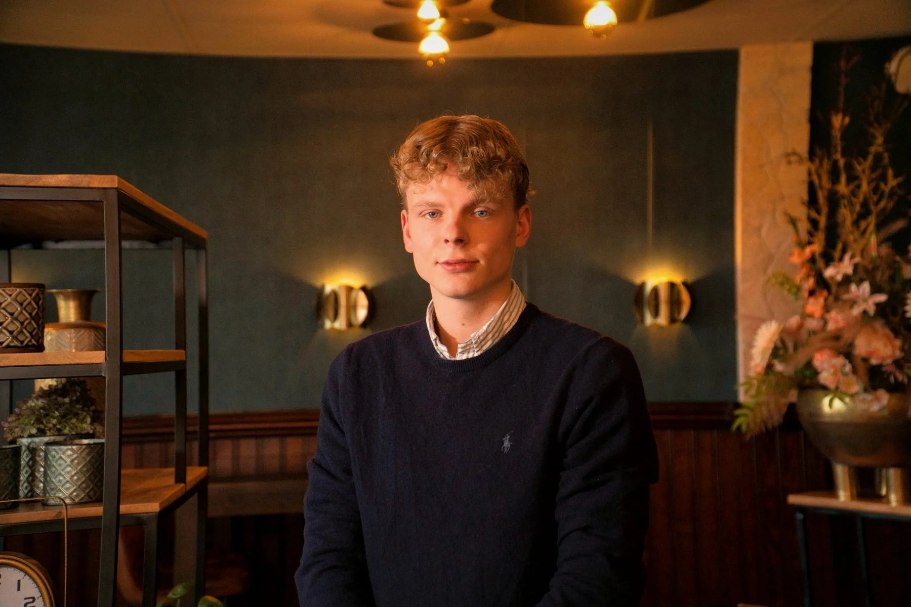 Een jonge man met blond, krullend haar en een lichte huid, zit in een sfeervolle, warm verlichte ruimte met bloemen aan de rechterkant en een staand rek met decoratieve items rechts. Hij draagt een donkerblauw trui en een blouse eronder.