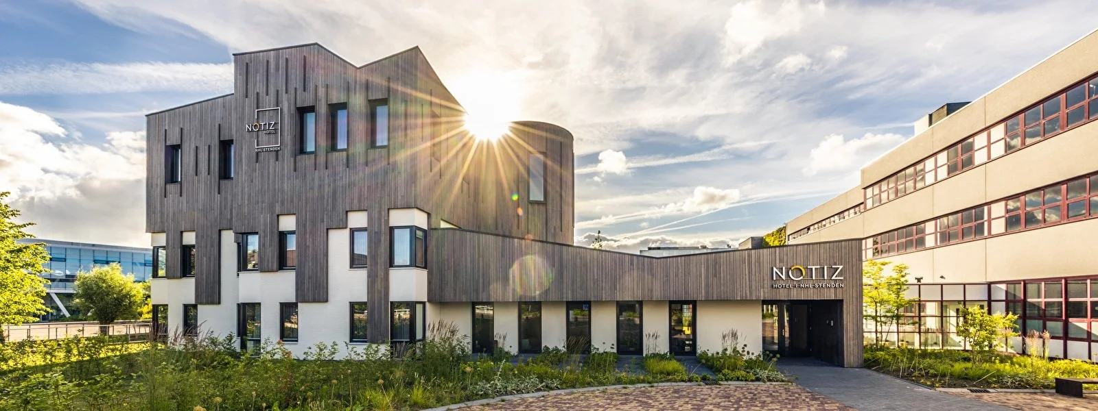 Moderne hotel met houten en witte gevel, zonsondergang en groene tuin eromheen.