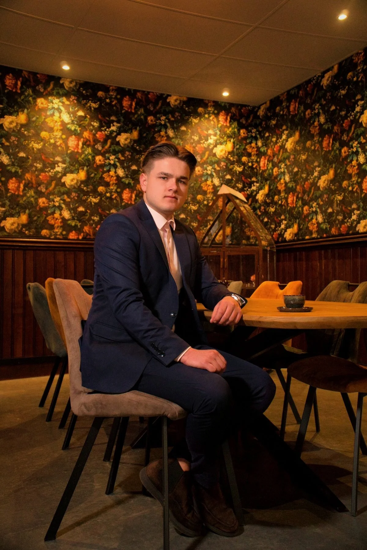 Een jonge man in een donkerblauw pak zit aan een houten tafel in een restaurant met een bloemrijke muur op de achtergrond.